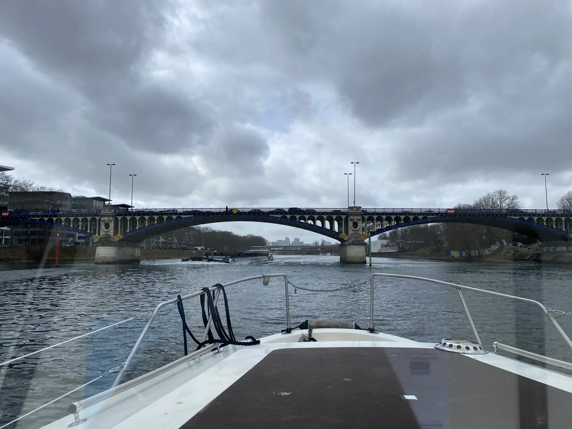 Étrave d'un bateau remontant la Marne vers un pont métallique de ville, berges aménagées de part et d'autre